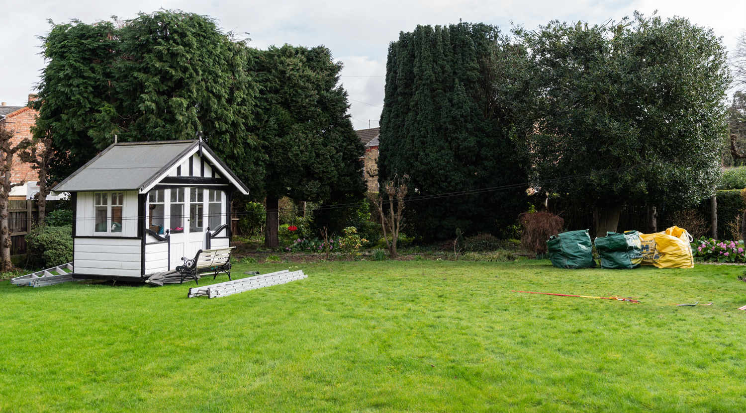 A cleared lawn with a white summerhouse and mature trees behind